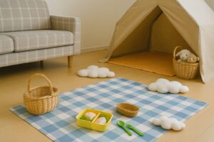 明るい部屋の中にブルーのチェック柄シートが敷かれ、お弁当風のランチセットとバスケットが並ぶ。奥にはテントとぬいぐるみが置かれ、室内ピクニックの雰囲気が広がる。